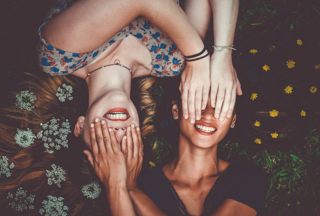Deux jeunes femmes allongées tête-bêche dans l’herbe fleurie, qui rient en se cachant les yeux avec les mains.